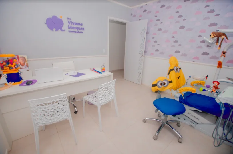 Consultório infantil da clínica: cadeira com capa de personagem, papel de parede de nuvens lilás e rosa e bichos de pelúcia
