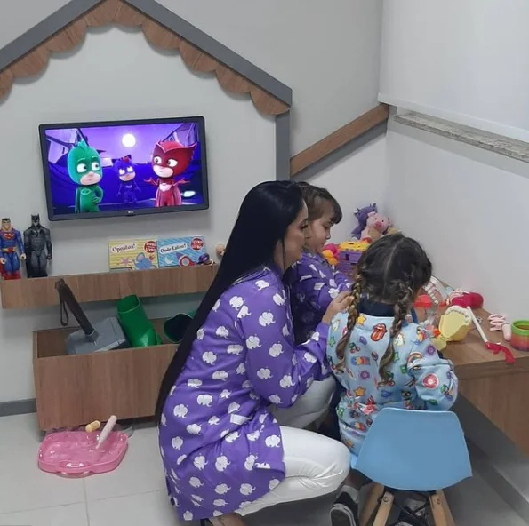 Espaço kids com TV na casinha, brinquedos e uma profissional da clínica brincando com uma criança de costas